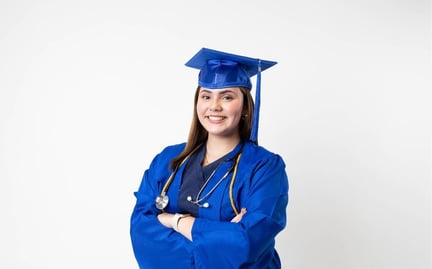 A student in a blue graduation cap and gown is smiling with arms crossed and wearing a stethoscope around neck