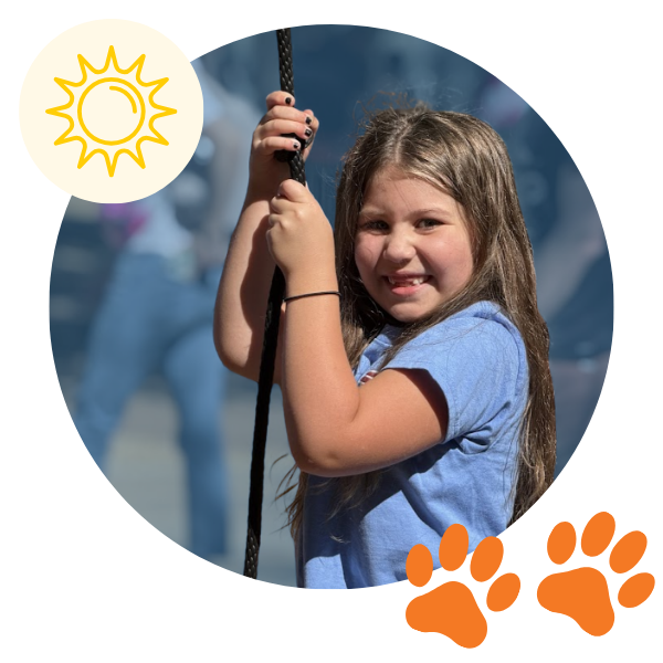A smiling elementary-aged girl wearing a blue shirt holds onto a playground rope, looking toward the camera during outdoor play on a sunny day.