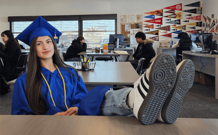 Grad student in a cap and gown, sitting at a table in a classroom, with her feet up on the desk
