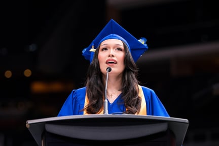 A high school graduate in a blue cap and gown speaks at a podium during a graduation ceremony. She is wearing a gold stole and tassel and appears proud and confident.