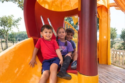 Three young children sit together smiling at the bottom of a yellow playground slide during outdoor playtime.