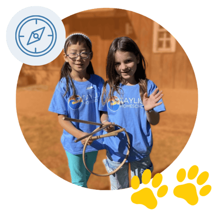 Two students wearing blue Taylion Homeschool shirts stand outdoors smiling and waving, holding ropes for an activity, shown inside an orange circle with a blue compass icon and yellow paw prints