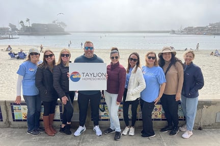 A group of staff stand together along a beach boardwalk, smiling and holding a Taylion Homeschool sign with the ocean, sand, and boats visible in the background.