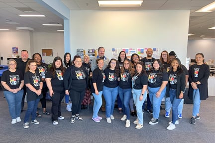 A large group of staff members stand together indoors, smiling and wearing matching black shirts, gathered in a shared office space.