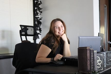 A smiling staff member sits at a desk in an office setting, resting her chin on her hand beside a laptop, computer monitor, and decorative books.