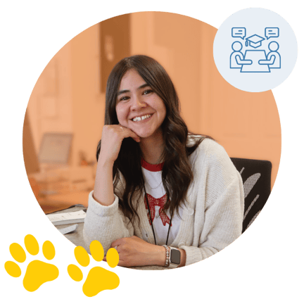 Smiling woman sitting at a desk in a classroom setting with a blue icon of two people at a table and two yellow paw prints, inside an orange-toned circular background.