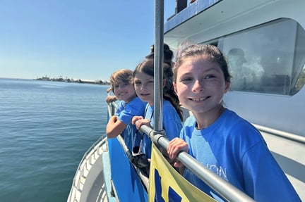 Three smiling students in blue shirts lean against the railing of a boat, looking out over the water with the ocean and distant shoreline visible on a sunny day.