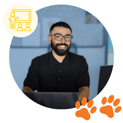 Smiling male teacher sitting at a desk with a laptop, featuring a yellow icon of a person teaching and two orange paw prints, inside a blue-toned circular background.