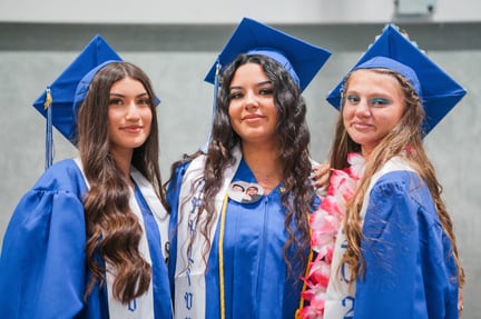Three female high school graduates wearing blue caps and gowns smile proudly, with white stoles and graduation accessories.
