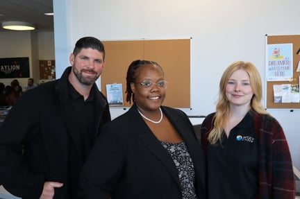 Three staff members stand together indoors, 1 man and 2 women are smiling at the camera in a school common area with bulletin boards visible behind them.