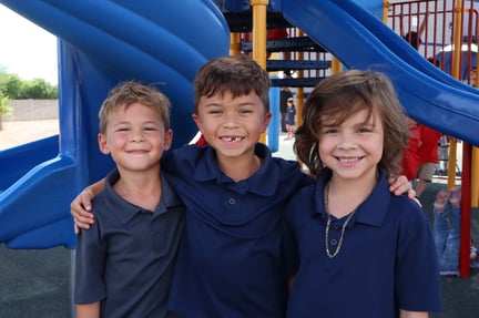 Three young boys wearing navy blue polo shirts smile with their arms around each other while standing in front of a blue playground slide.
