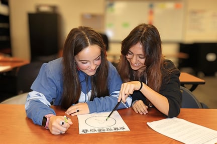 Two students sit together at a table, smiling and working on a paper as one of them points to the page with a pencil.