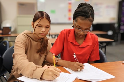 Two students sit side by side at a table, focused as they write on worksheets with pencils during a classroom activity.